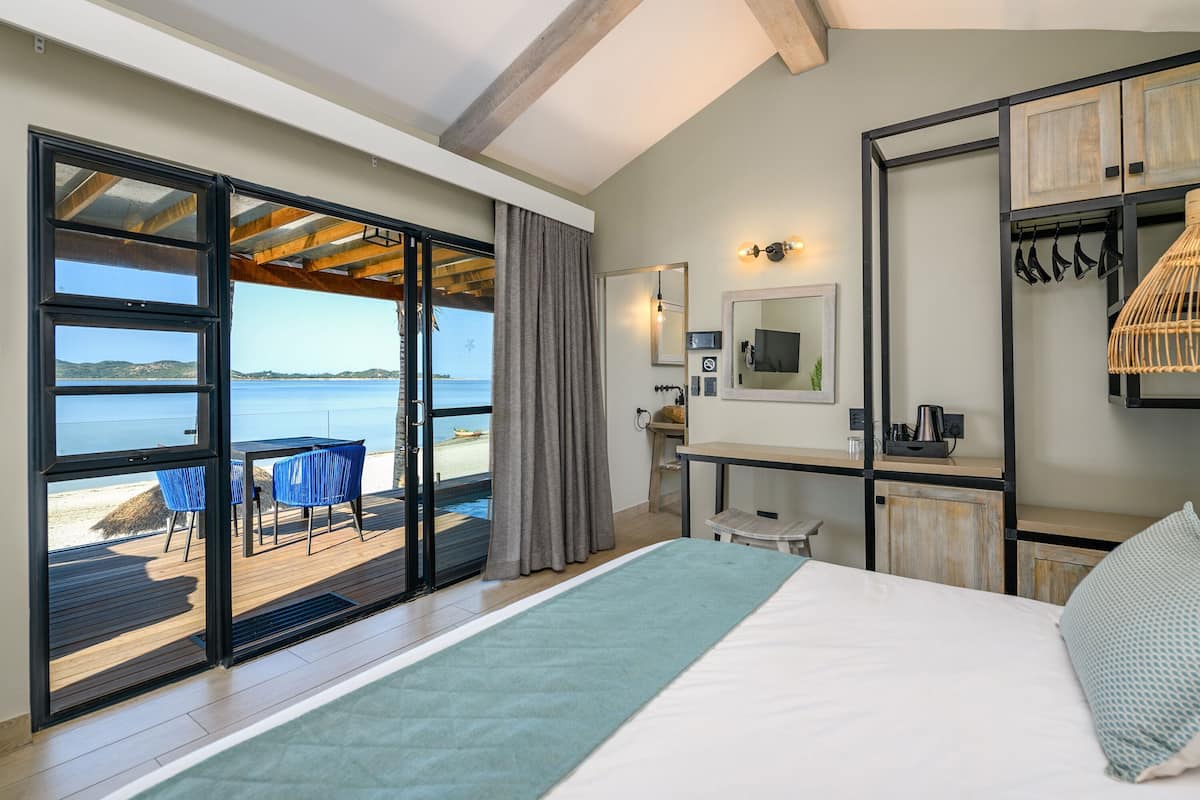 A bedroom with a bed, a desk, a chair, a mirror, and a view of the sea through a sliding door.