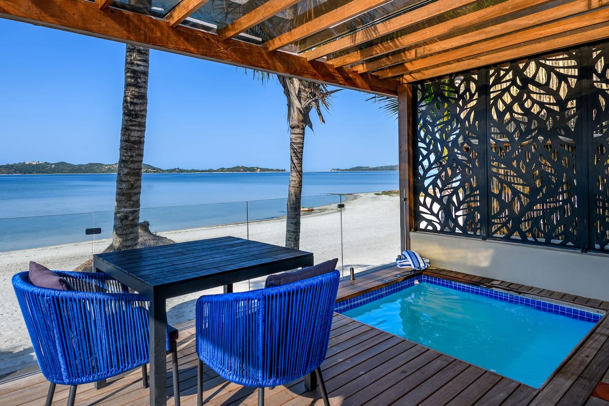 A pool area with a wooden deck, two blue chairs, a table, and a view of the beach and sea.