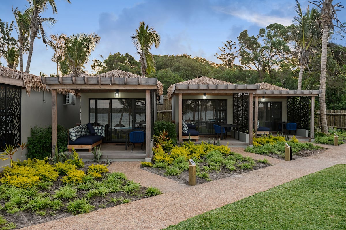 A resort with two thatched-roof structures, each with a seating area and a glass door.