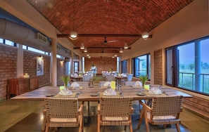 A spacious dining area with a high ceiling and brick walls, featuring large windows and a view of the outdoors. - Wagoba Eco lodge (Bhadravathi)