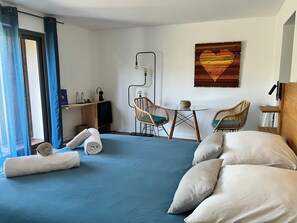 Um quarto com uma cama, duas cadeiras de vime, uma mesinha, uma luminária na parede e um quadro.