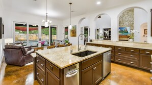 A spacious kitchen with granite countertops, stainless steel appliances, and a large island. There is a dining area with a leather sofa and a chandelier. The kitchen has a stone accent wall and large windows with a view of a green landscape.