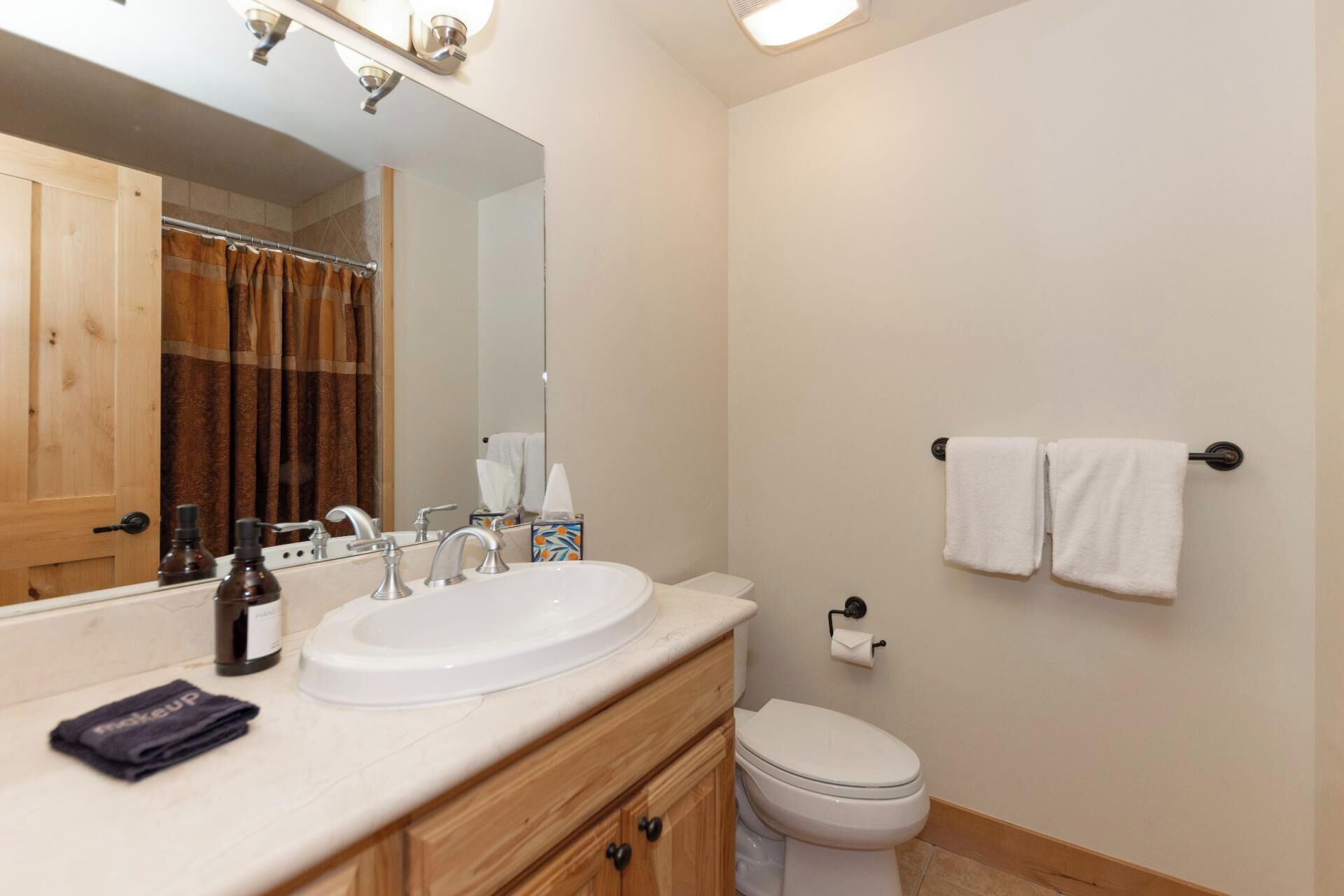 A bathroom with a sink, mirror, and toilet. There are towels hanging on the wall and a bottle of hand soap on the counter.