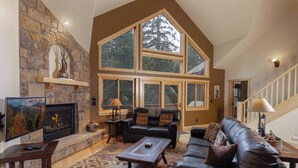 A spacious living room with a stone fireplace, large windows, and a high ceiling.