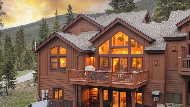 A two-story wooden house with a balcony, a hot tub, and a view of the surrounding forest.