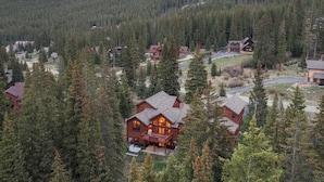 A large log cabin with a deck and hot tub, surrounded by dense forest and mountains.