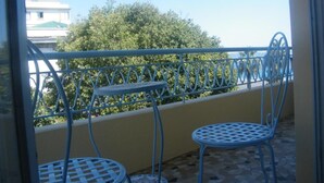 Ein Balkon mit zwei Metalltischen und -stühlen, einer gelben Wand und Blick auf Bäume und Gebäude.