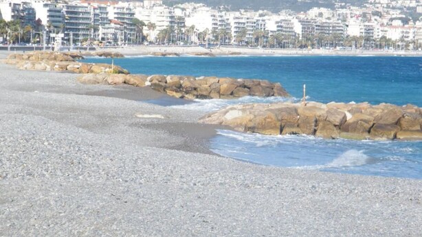 Ein Kiesstrand mit einem felsigen Wellenbrecher und einer Stadtkulisse im Hintergrund.