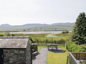 A stone building with a grassy yard, a picnic table, and a view of a lake and hills.