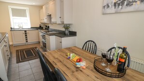 A kitchen with white cabinets, a wooden dining table, and a black chair. There is a bottle of wine and a glass on the table. - Minffordd (Blaenau Ffestiniog)