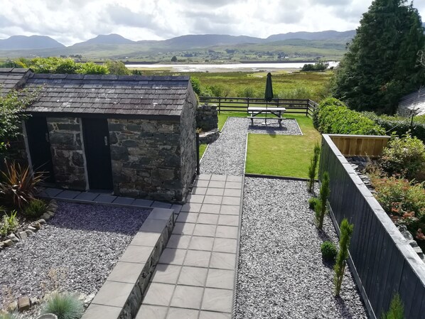 A stone building with a small patio area, a grassy lawn, and a view of a river and mountains in the distance.