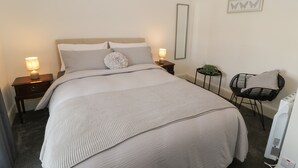 A neatly made bed with a grey and white color scheme, a bedside table with a lamp, a small round table with a plant, and a black wicker chair. - Minffordd (Blaenau Ffestiniog)