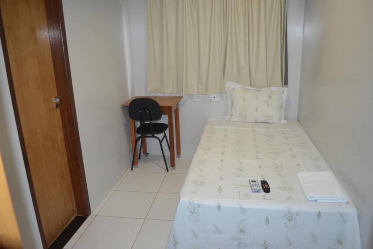 A small, single-bed room with a desk and chair, a window with curtains, and a door.