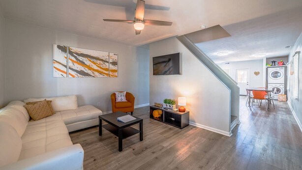 A modern living room with a white sofa, a coffee table, and a ceiling fan. There is a staircase leading to a kitchen area with a washing machine.