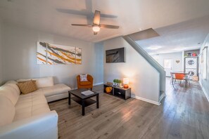 A modern living room with a white sofa, a coffee table, and a ceiling fan. There is a staircase leading to a kitchen area with a washing machine. - Orange Crush Townhome I, Beach & Pet-Friendly (Cape Charles)