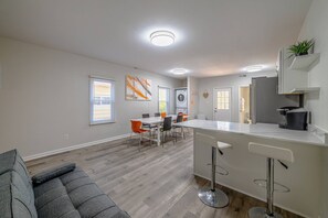 Una cocina moderna con una isla central, una zona de comedor con mesa y sillas, y un sofá gris en un amplio salón.