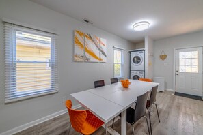 A dining area with a white table, orange chairs, and a vase of orange flowers. There are two windows with blinds, a washing machine, and a door with a doormat. - Orange Crush Townhome II, Beach & Pet-Friendly (Cape Charles)