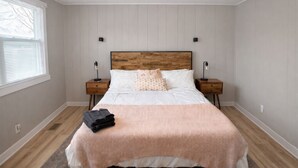Camera da letto con testiera in legno, biancheria bianca, due comodini con lampade e una finestra con persiane.