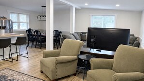 Un soggiorno moderno con una TV a schermo piatto, poltrone beige, un'area pranzo con un tavolo e sedie in legno, e un angolo cottura con frigorifero e macchina del caffè.