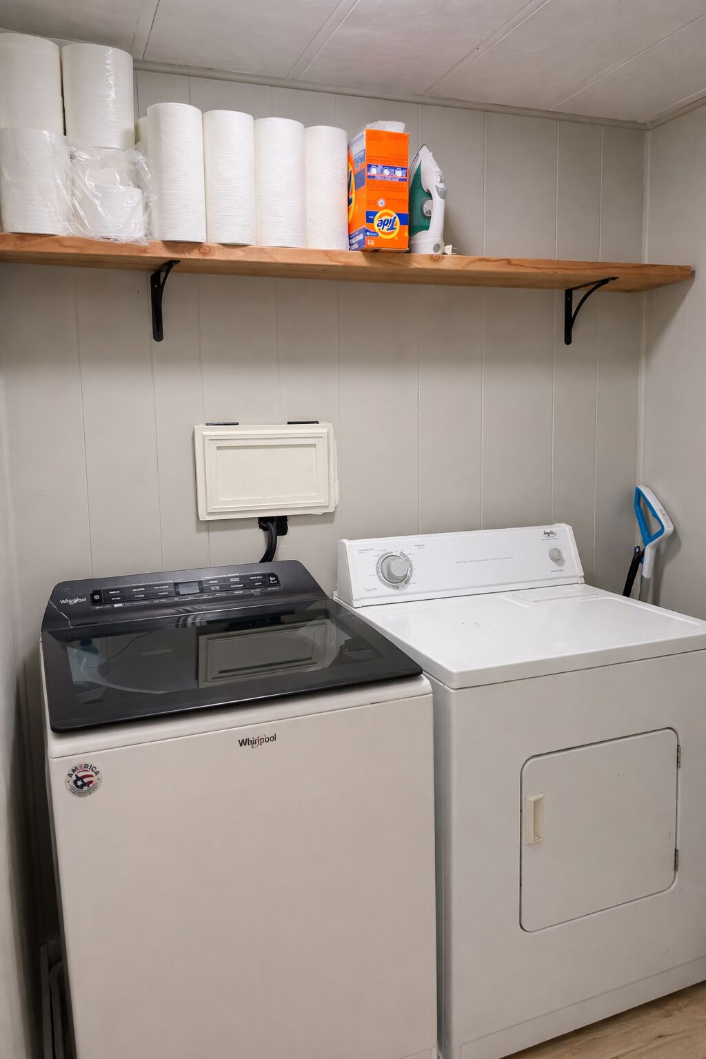 Un espace buanderie équipé d’un lave-linge et d’un sèche-linge Whirlpool, d’une étagère avec des essuie-tout et d’une boîte de lessive, ainsi que d’un panneau de commande fixé au mur.
