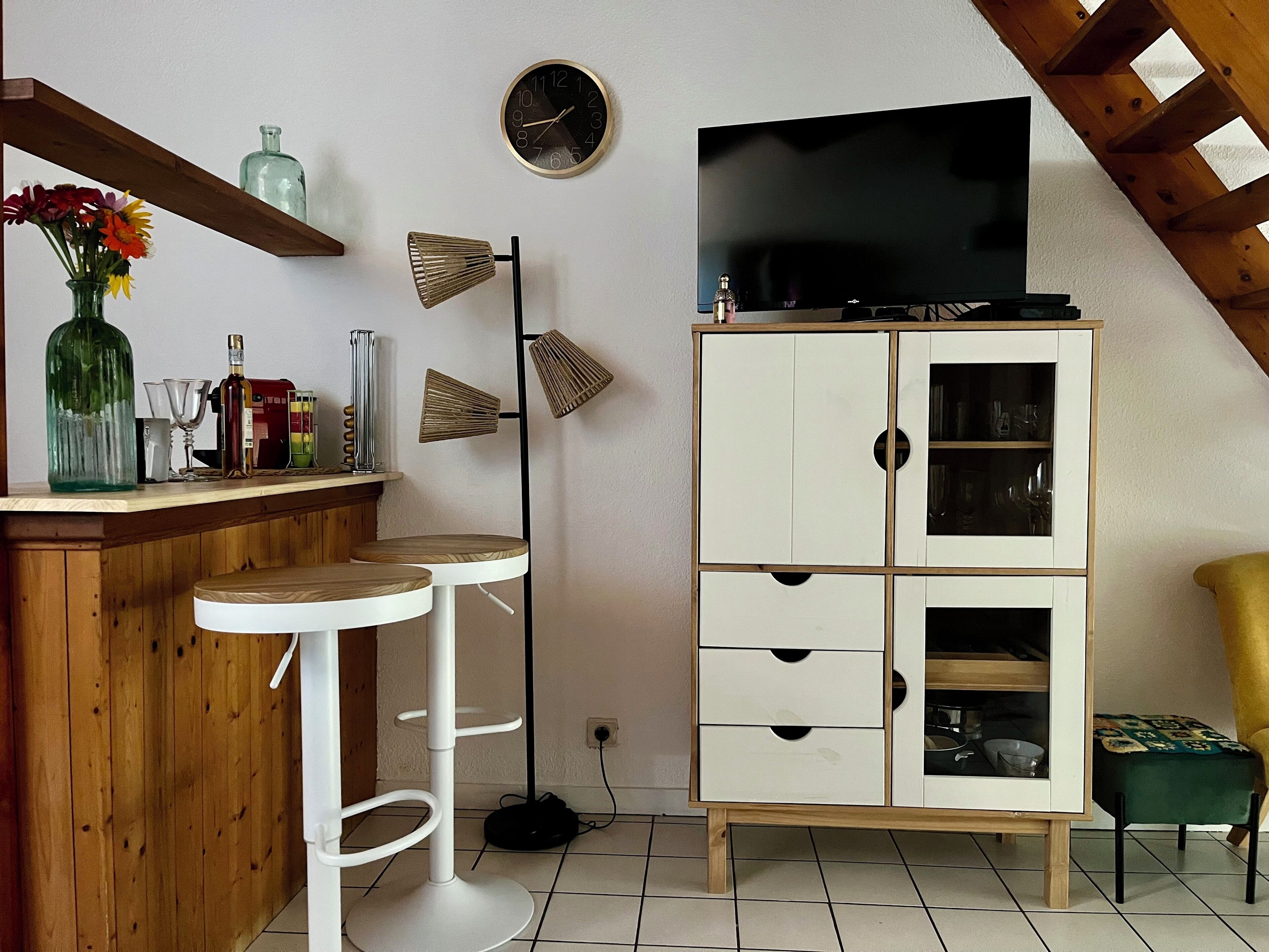 A room with a TV on a cabinet, a bar area with stools, a clock on the wall, and a wooden ceiling.