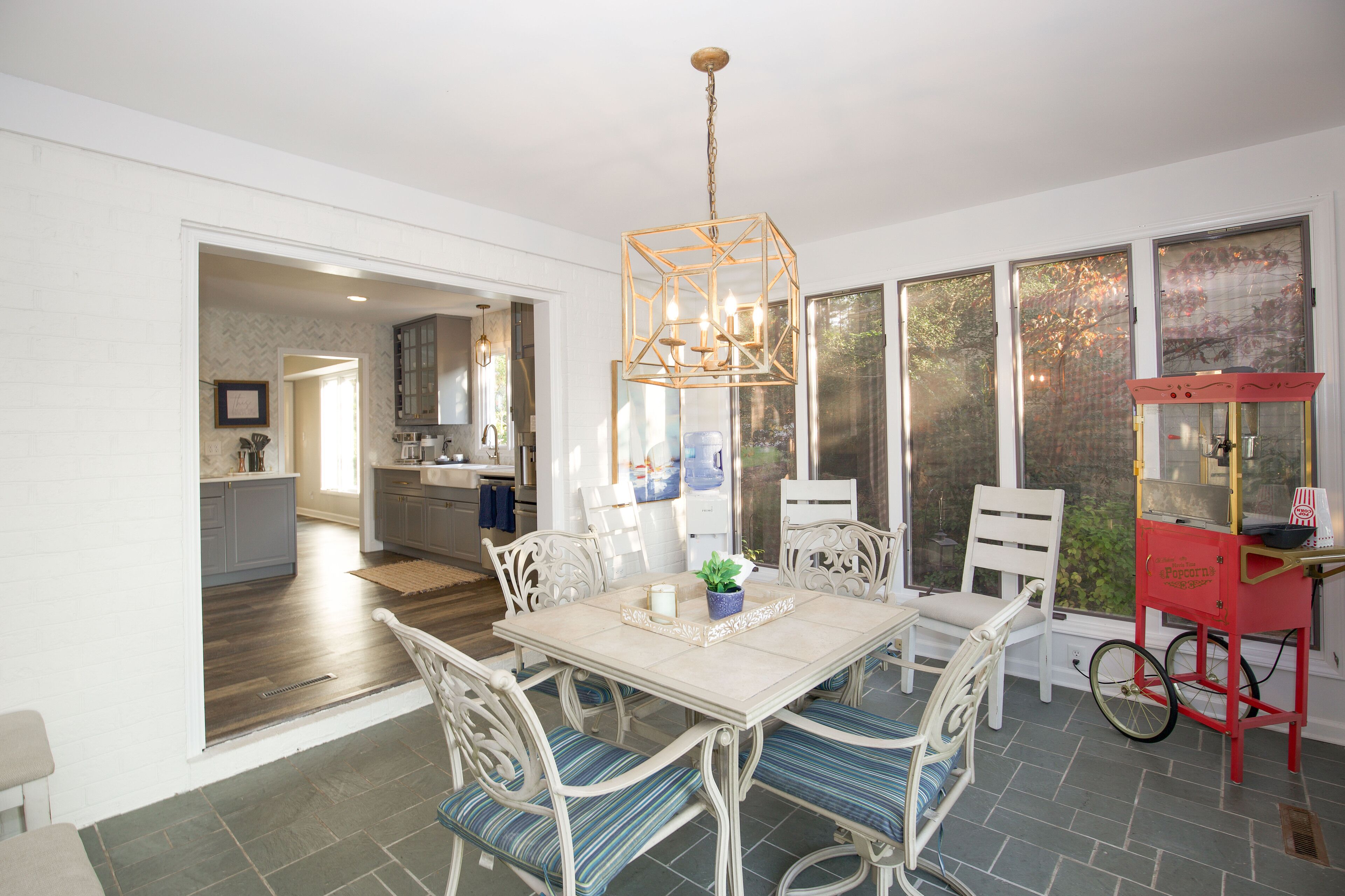 A dining area with a table set for four, surrounded by white chairs with blue cushions. A red cart with a vintage design is placed near the window.
