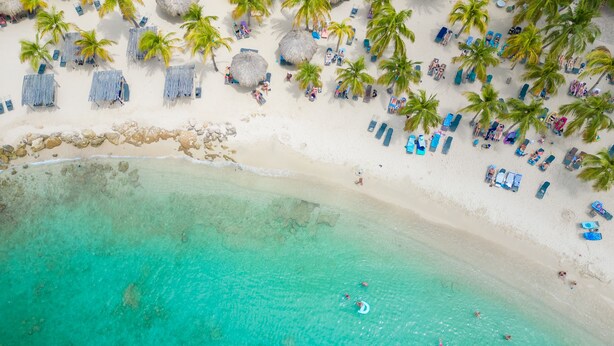 An aerial view of a beach with clear turquoise water, palm trees, and beach chairs. - Beachfront, sea view 3 BDR luxury The Shore @ Blue Bay Beach and Golf Resort (Willemstad)