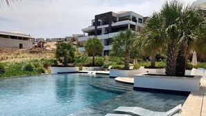 A swimming pool with lounge chairs, palm trees, and modern buildings in the background. - Beachfront, sea view 3 BDR luxury The Shore @ Blue Bay Beach and Golf Resort (Willemstad)