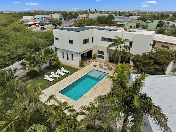 A modern two-story building with a swimming pool, surrounded by palm trees and a view of a town.