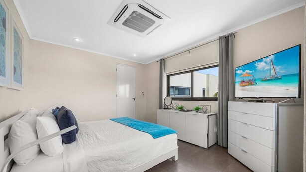 A hotel room with a bed, a desk, a television, and a window with a view of the beach. - Villa Haven: 6BR w/ Private Pool Near Palm Beach! (Noord)