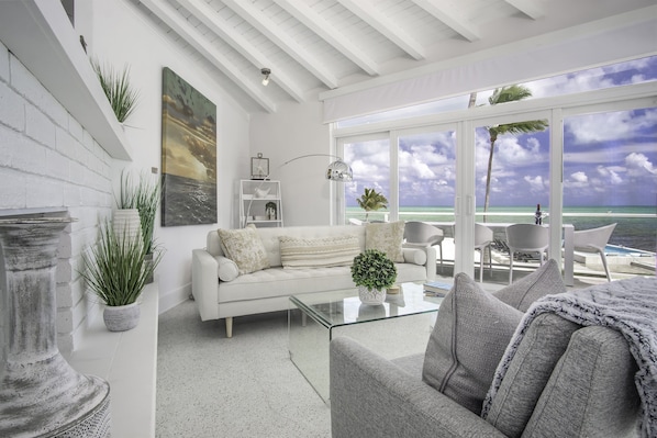 A modern living room with a white sofa, a glass coffee table, and a view of the ocean through large windows. - Oceanfront 2 Homes | Private Beach & Pool | 3 Acre (Islamorada)