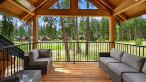 A covered outdoor deck with wooden flooring, wicker furniture, and a view of a golf course and surrounding trees.