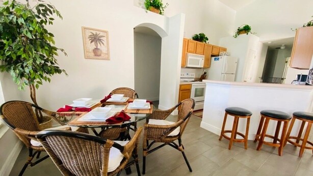 Un comedor con sillas de mimbre y una mesa preparada para cuatro, una cocina con armarios de madera y un microondas, y una pequeña zona de bar con taburetes.