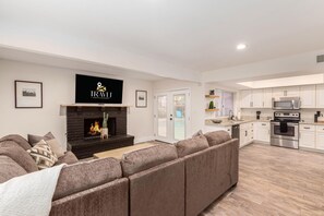A modern living room with a fireplace, a sofa, a television, and a kitchen area with appliances. - Obispo Modern Oasis with Heated Pool and BBQ (Mesa)
