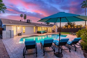 A swimming pool with lounge chairs, an umbrella, and a house with a patio.