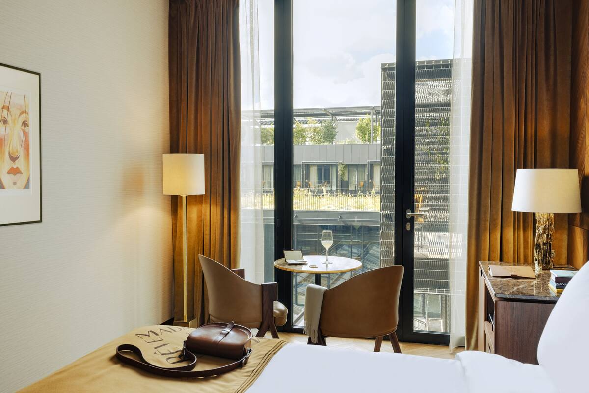 A hotel room with a bed, two chairs, a table with a laptop and a glass of wine, a handbag, and a view of a building through the window.