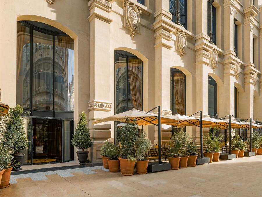 Exterior de un hotel con amplias ventanas arqueadas, mobiliario exterior con sombrillas y plantas en macetas.