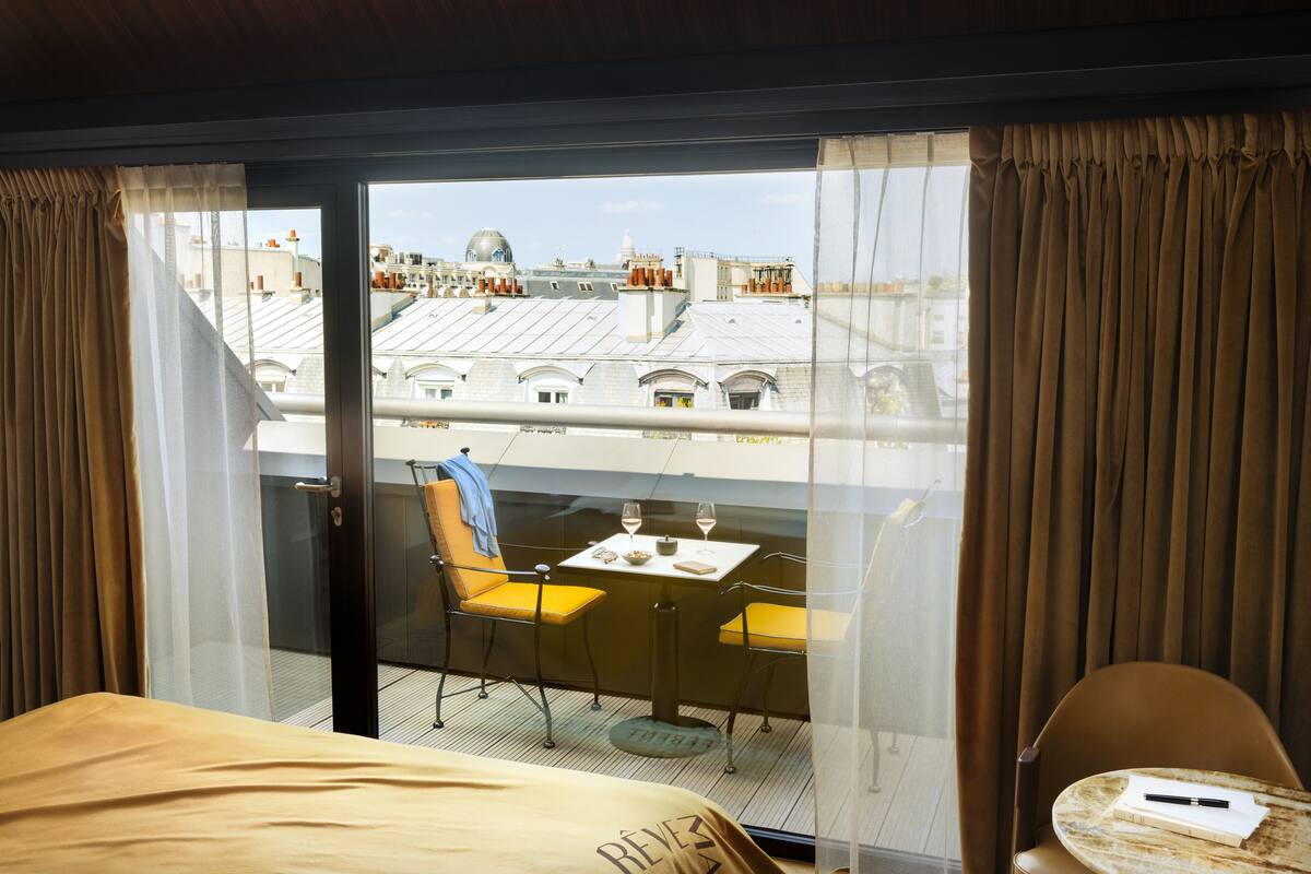 A hotel room with a balcony, a bed, a table with two glasses of wine, and a chair.