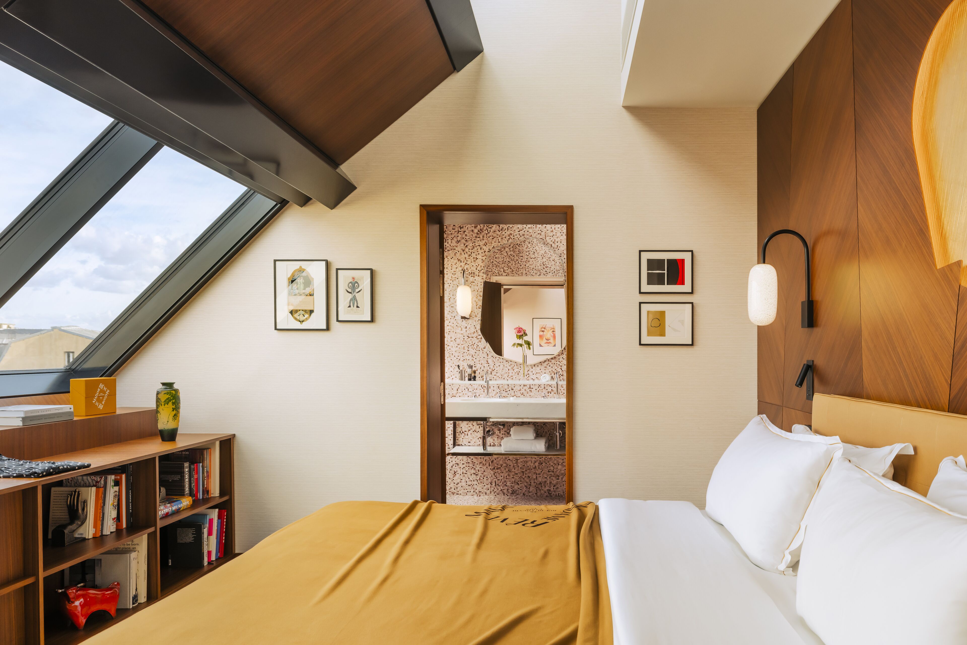 A bedroom with a skylight, a bed with white linens, a wooden headboard, a bookshelf, and a bathroom visible through an open door.