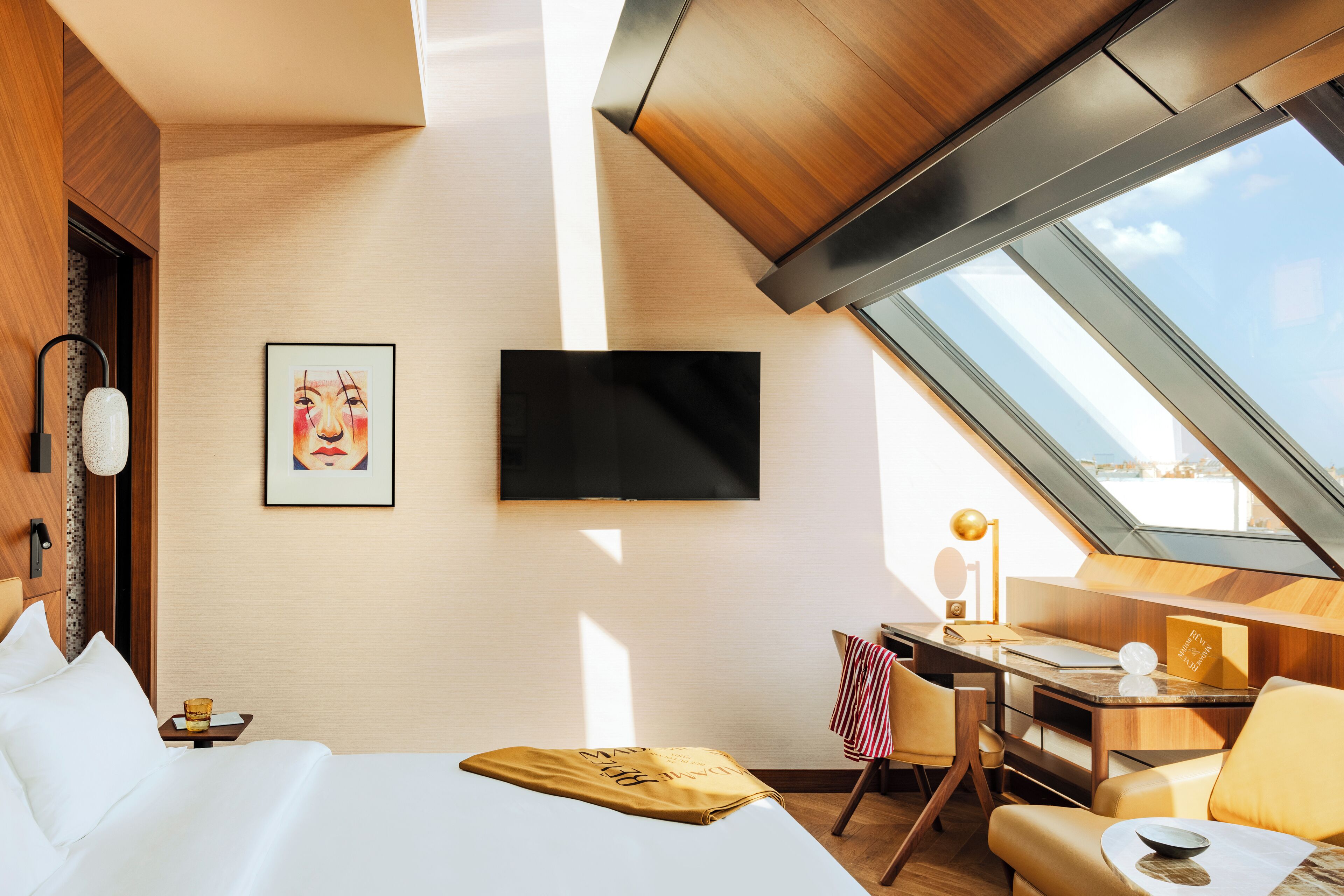 A modern hotel room with a bed, a desk, a chair, a TV, and a skylight.