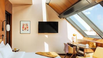 A modern hotel room with a bed, a desk, a chair, a TV, and a skylight.