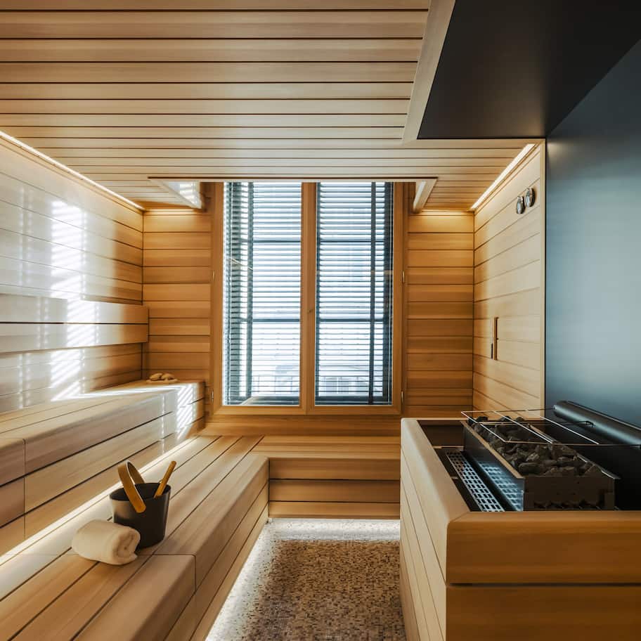 A sauna room with wooden walls, a bench, a bench with a towel, a small table with a towel and a brush, and a window with blinds.
