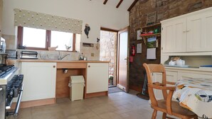 A kitchen with a stove, sink, and wooden cabinets. There is a window with a patterned valance and a door leading to another room. - The Proven House (Preston)