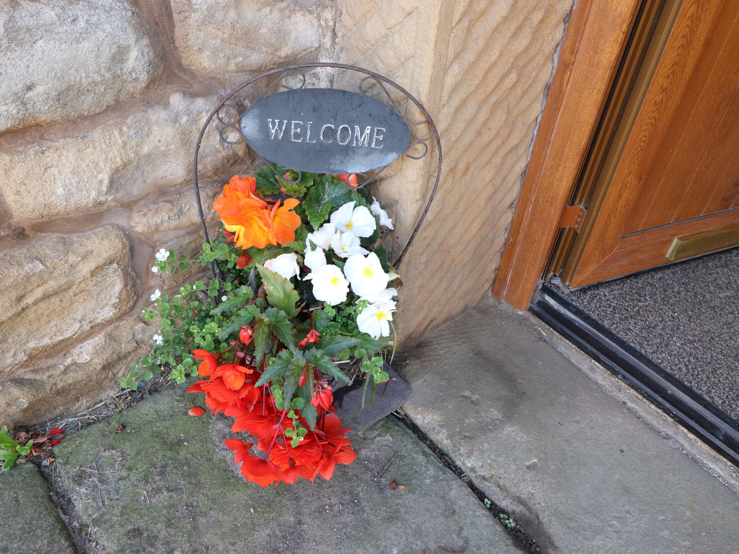 A welcome sign with flowers in front of a stone wall and a wooden door.