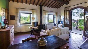 A cozy living room with a wooden table, a sofa, and a TV. There are windows with curtains, a fireplace, and a view of greenery outside. - Le Rose di Montagnana (San Gimignano)