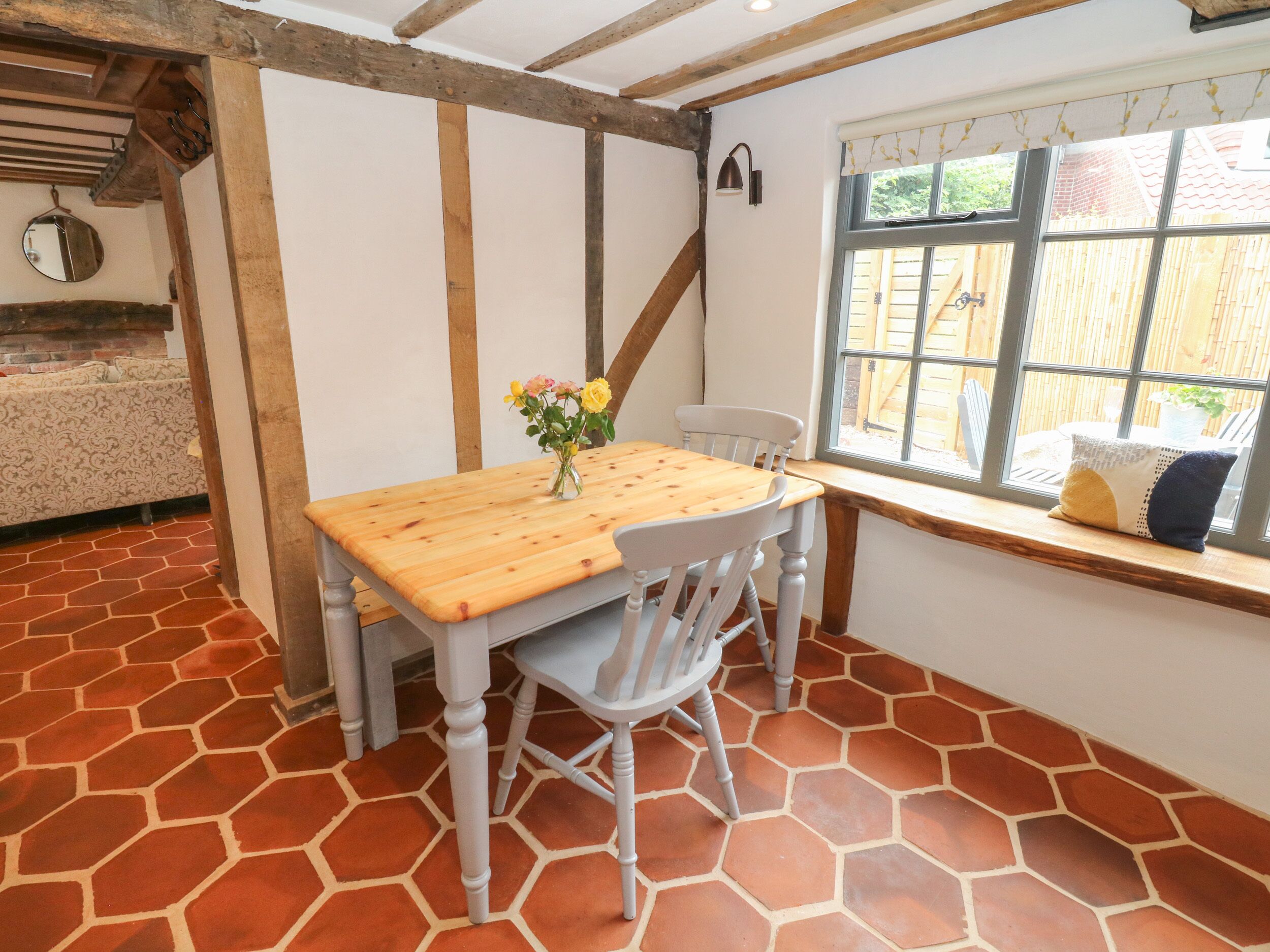 A dining area with a wooden table and chairs, a vase of flowers, and a window with a view of a wooden fence.