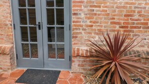 Unas puertas dobles grises con paneles de vidrio, un muro de ladrillo y una planta en maceta con hojas largas y puntiagudas.