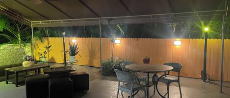 An outdoor patio area with a covered seating area, including a round table and chairs, surrounded by potted plants and a wooden fence.