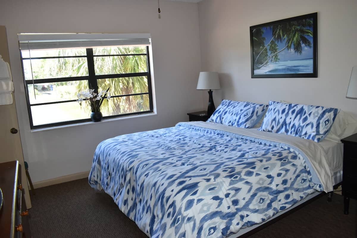 A bedroom with a bed, a window with a view of palm trees, a nightstand with a lamp, and a painting of a beach scene.