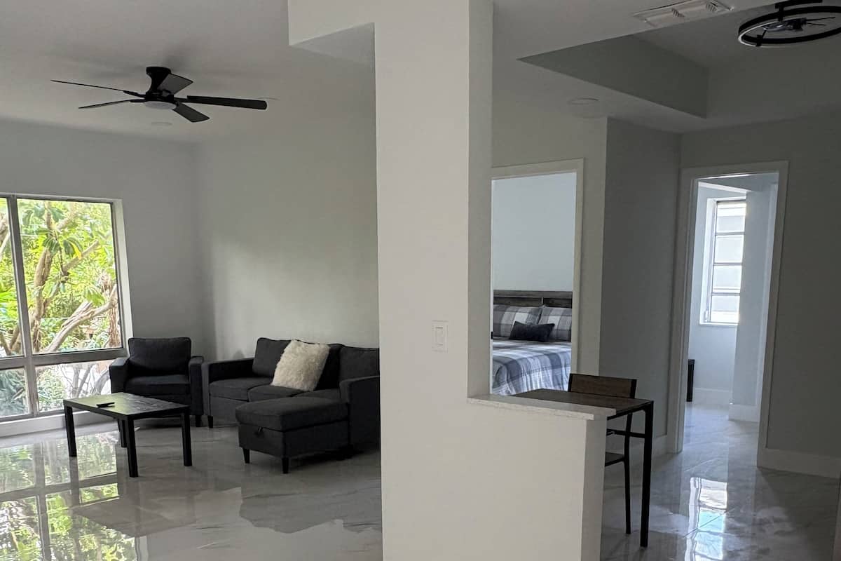 A spacious living room with a ceiling fan, a sofa, a coffee table, and a view of a bedroom through an open doorway.